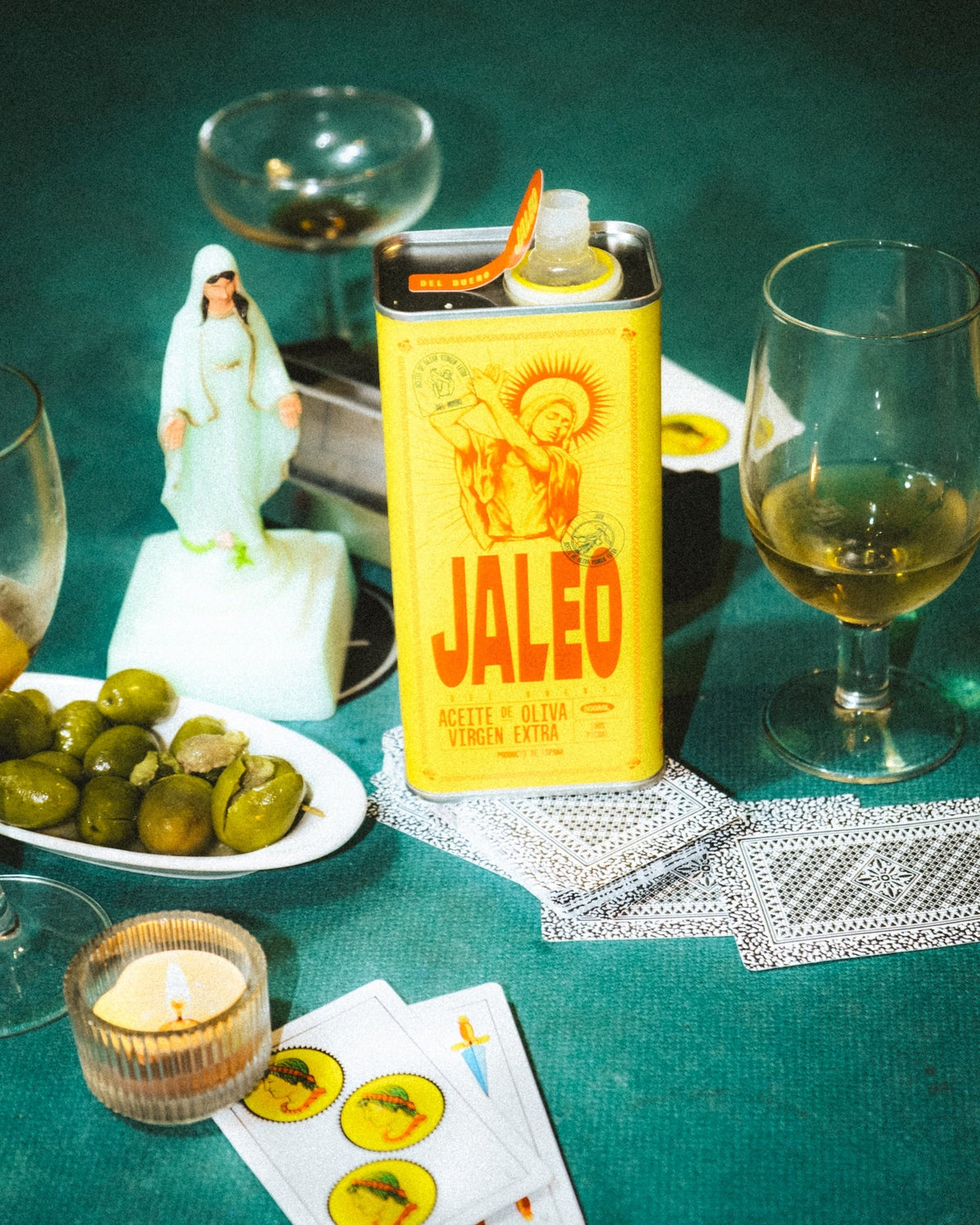 Bodegón sobre fondo verde con la lata de Jaleo del bueno, copas de vino, aceitunas y una figura de cerámica